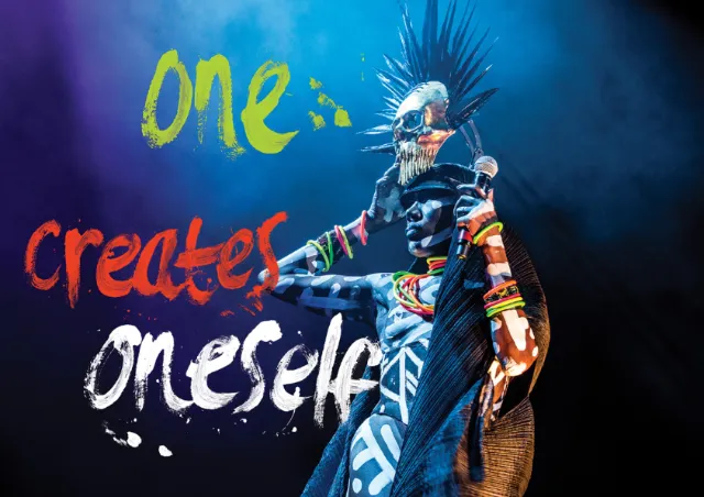 Performer in tribal attire and spiked headdress on stage with colorful text