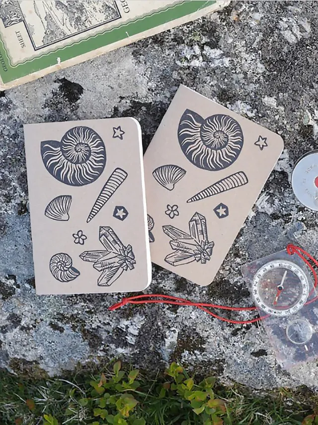 Two notebooks with fossil linocut covers on rock with compass and map by Becca Thorne
